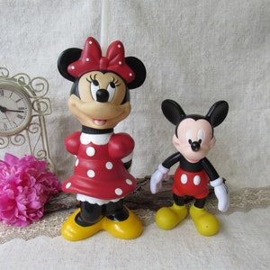 Puede incluir: Dos figuras de personajes de Disney: Minnie Mouse y Mickey Mouse. Minnie Mouse viste un vestido rojo con lunares blancos, un lazo a juego y zapatos amarillos. Mickey Mouse lleva pantalones cortos rojos, guantes blancos y zapatos amarillos. Un reloj decorativo y flores rosas en el fondo.