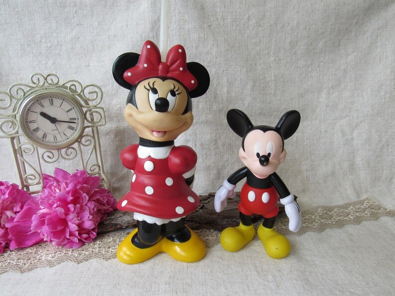 Puede incluir: Dos figuras de personajes de Disney: Minnie Mouse y Mickey Mouse. Minnie Mouse viste un vestido rojo con lunares blancos, un lazo a juego y zapatos amarillos. Mickey Mouse lleva pantalones cortos rojos, guantes blancos y zapatos amarillos. Un reloj decorativo y flores rosas en el fondo.