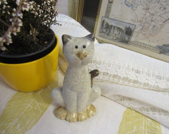 May include: A ceramic cat figurine with a gray and white speckled finish. The cat is sitting with its tail curled around its legs. The figurine has a whimsical, cartoonish style.