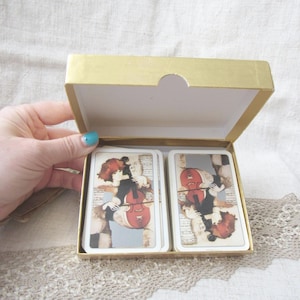 May include: A gold-coloured box containing a deck of playing cards. Each card displays a vintage illustration of a person playing a violin. The cards have a cream-coloured background with musical notes.
