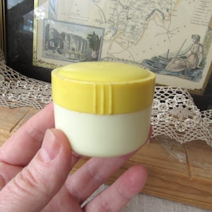 Könnte beinhalten: Ein kleiner, zweifarbiger Behälter mit gelbem Deckel und cremefarbenem Boden. Der Deckel hat vertikale Rillen. Der Behälter wird in einer Hand gehalten, mit einer Vintage-Karte und Kunstwerken im Hintergrund.