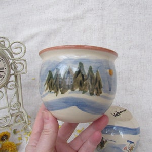 May include: A white ceramic mug with a blue and green winter scene painted on it. The mug has a brown rim and a handwritten inscription on the bottom that reads "Valonia".