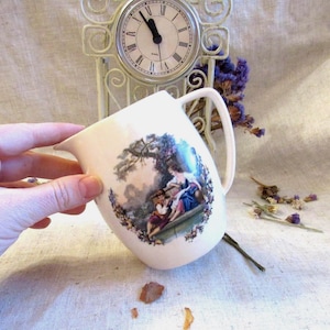 Może przedstawiać: Kremowy ceramiczny dzbanek z uchwytem, przedstawiający sielankową scenę z postaciami w ogrodzie. Dzbanek ma zakrzywioną wylewkę i biały uchwyt. Z tyłu znajduje się zegar w stylu vintage.