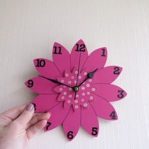 Puede incluir: Un reloj de pared rosa con forma de flor con números y manecillas negras. El reloj tiene un centro decorativo con detalles de purpurina plateada. El reloj tiene aproximadamente 30 cm de diámetro.