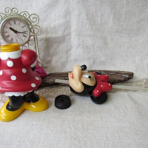 Puede incluir: Una figura de Minnie Mouse, vestida con un vestido rojo con lunares blancos, zapatos negros y suelas amarillas. Una tapa de botella negra est&aacute; al lado. Un reloj decorativo y flores rosas est&aacute;n en el fondo.