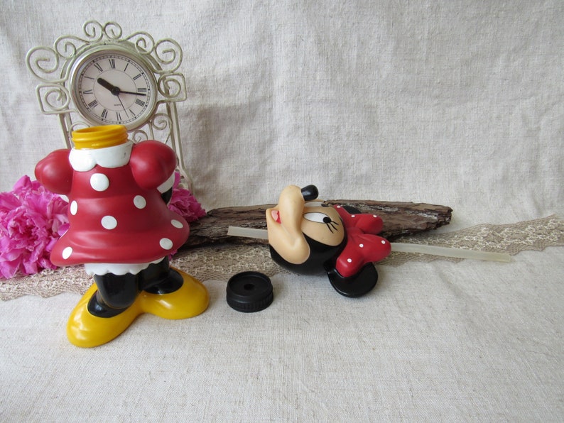 Puede incluir: Una figura de Minnie Mouse, vestida con un vestido rojo con lunares blancos, zapatos negros y suelas amarillas. Una tapa de botella negra est&aacute; al lado. Un reloj decorativo y flores rosas est&aacute;n en el fondo.