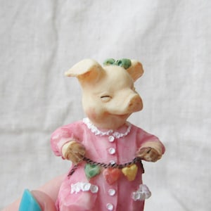 Op de afbeelding: Een roze varkensfiguur dat een roze jurk draagt met een witte kantversiering en een ketting van hartjes om zijn nek. Het varken heeft een groene strik op zijn kop.