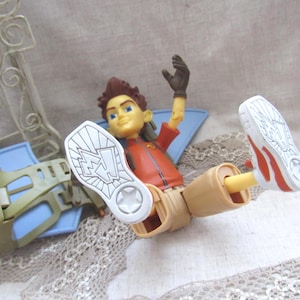May include: A toy action figure with brown hair, an orange jacket, and tan pants. The figure is lying down with one arm raised and the soles of its white shoes with red accents facing the viewer. A blue and green accessory is in the background.
