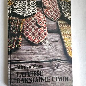 May include: A hardback book titled Mirdza Slava LATVIEŠU RAKSTAINIE CIMDI featuring a cover image of several colourful, intricately patterned knitted mittens arranged on a weathered wooden surface with visible grain and knots.