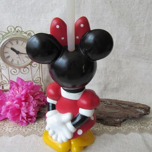Puede incluir: Un candelabro de Minnie Mouse. La cabeza negra tiene orejas grandes y un lazo rojo con lunares blancos. El vestido rojo tiene lunares blancos, guantes blancos y zapatos amarillos. Una vela blanca est&aacute; insertada en la cabeza.