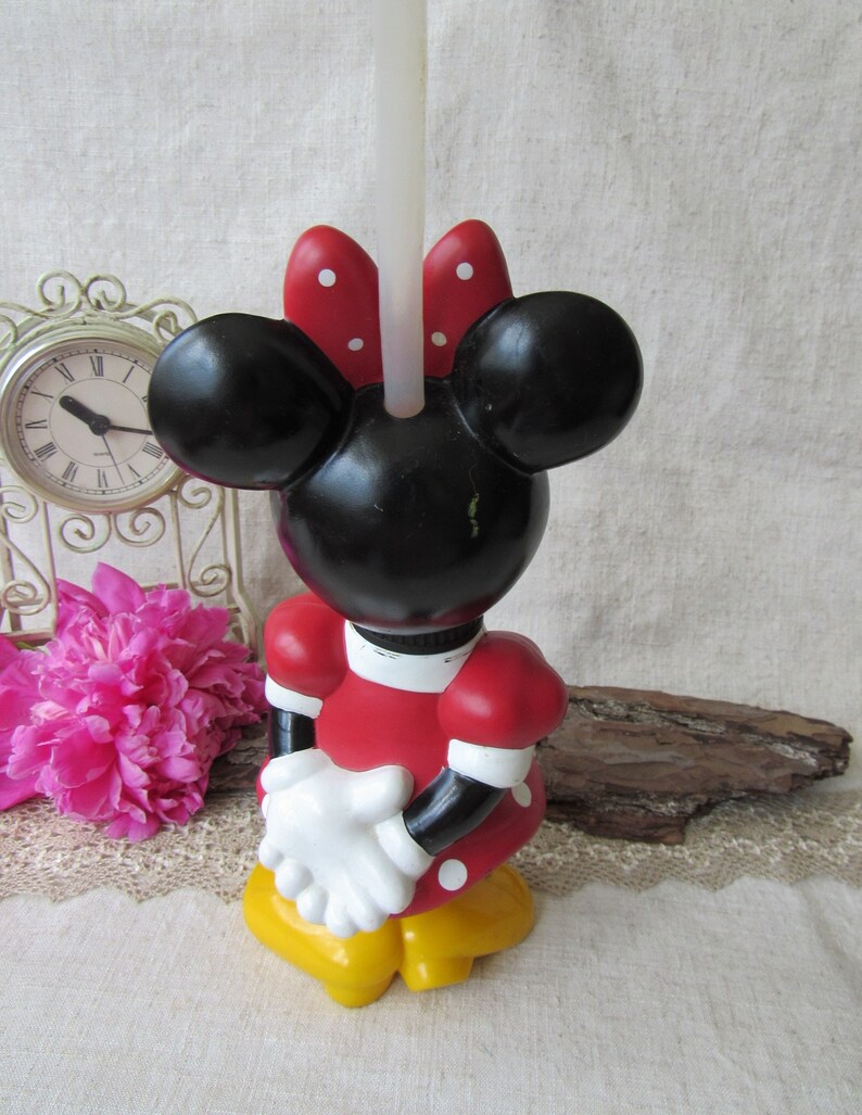Puede incluir: Un candelabro de Minnie Mouse. La cabeza negra tiene orejas grandes y un lazo rojo con lunares blancos. El vestido rojo tiene lunares blancos, guantes blancos y zapatos amarillos. Una vela blanca est&aacute; insertada en la cabeza.