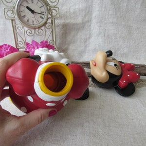 Puede incluir: Un juguete de Minnie Mouse rojo con lunares blancos y una abertura amarilla y blanca. El juguete est&aacute; sobre una superficie de color claro. Tambi&eacute;n se ve una segunda figura de Minnie Mouse.