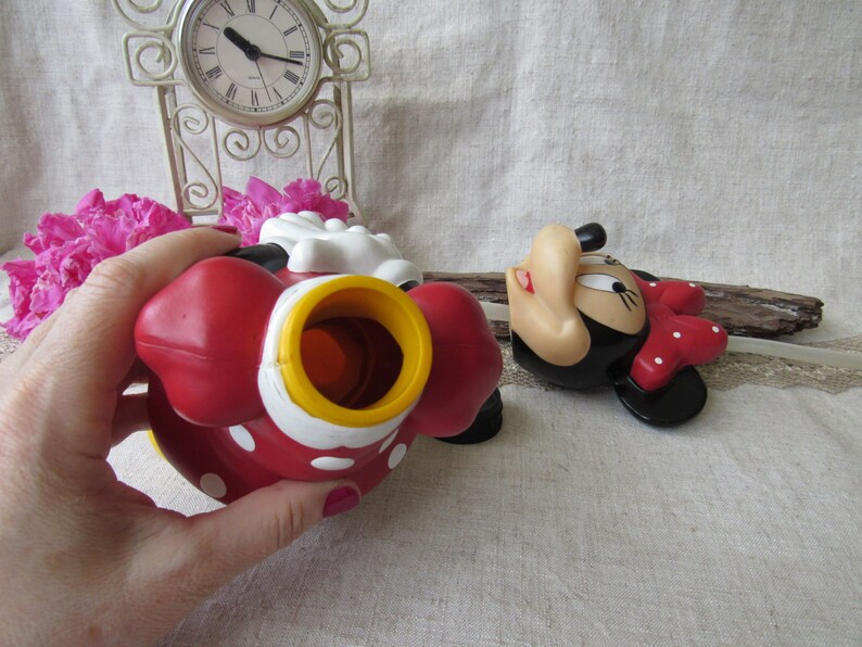 Puede incluir: Un juguete de Minnie Mouse rojo con lunares blancos y una abertura amarilla y blanca. El juguete est&aacute; sobre una superficie de color claro. Tambi&eacute;n se ve una segunda figura de Minnie Mouse.