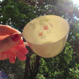 Op de afbeelding: Een wit theekopje met een bloemenpatroon, vastgehouden in een hand met rode nagellak, tegen een achtergrond van groene bomen en een heldere, zonnige lucht.