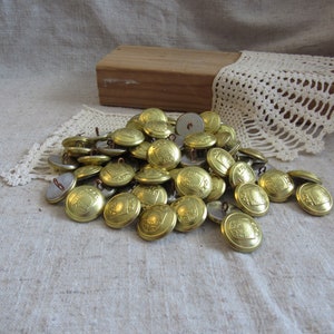 May include: A pile of vintage brass buttons with a shield design. The buttons are scattered on a beige linen surface, with a wooden block and a piece of lace in the background. The buttons have a shiny, metallic finish.