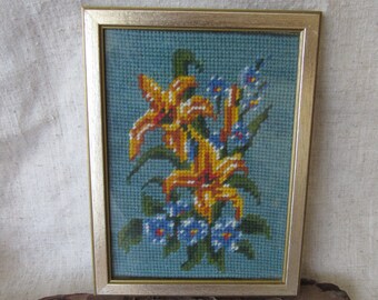 Puede incluir: Un bordado en punto de cruz enmarcado que presenta un arreglo floral con lirios amarillos y naranjas vibrantes y pequeñas flores azules sobre un fondo verde azulado, dentro de un marco rectangular de metal dorado.