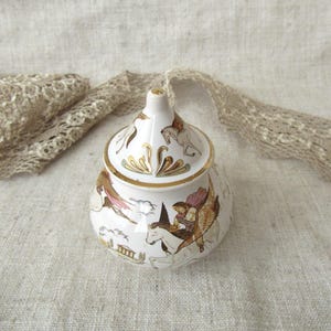 Può includere: Un barattolo in ceramica bianca con coperchio, bordi dorati e una scena dipinta raffigurante una figura a cavallo di un cavallo alato. Il coperchio appuntito è decorato con motivi dorati, mentre la base mostra un paesaggio architettonico.