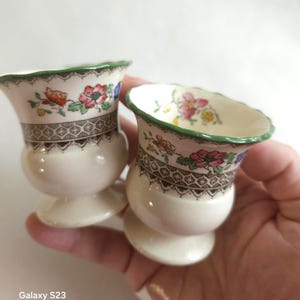 Puede incluir: Dos hueveras de cerámica de color crema con un borde festoneado verde y un patrón floral con flores rosas, naranjas y azules. Una banda geométrica marrón rodea la sección superior de cada huevera, que se asienta sobre una base circular.