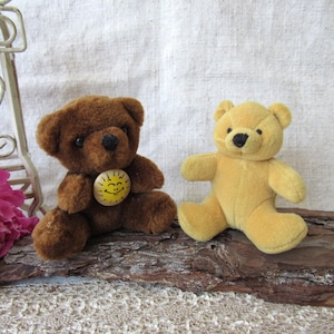 May include: Two small teddy bears. One is brown with a yellow button featuring a smiling sun. The other is yellow. Both are plush toys with black eyes and noses. They are sitting on a piece of wood.