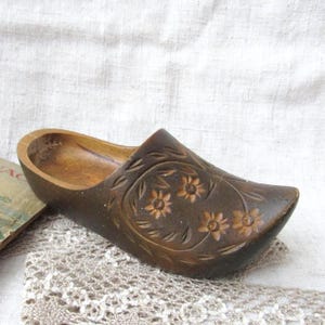 Puede incluir: Un zueco de madera marrón con detalles florales tallados. El zapato presenta un interior hueco y descansa sobre un tapete de encaje. La superficie del zueco tiene un aspecto oscuro y envejecido.