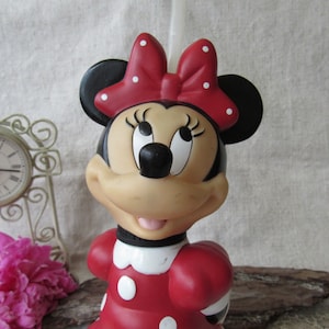 Puede incluir: Figura de Minnie Mouse, con un vestido rojo con lunares blancos y un lazo a juego. La figura tiene orejas negras, una nariz negra y una expresi&oacute;n sonriente. Un art&iacute;culo de colecci&oacute;n.