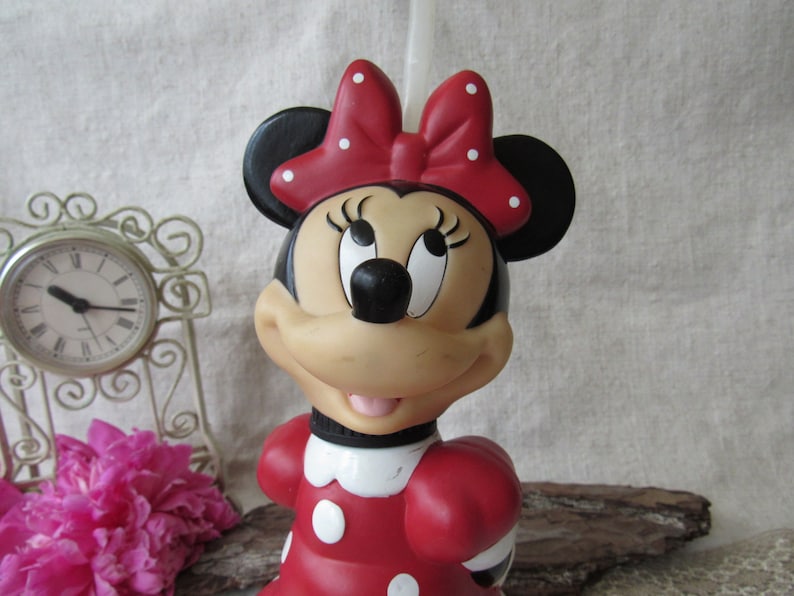 Puede incluir: Figura de Minnie Mouse, con un vestido rojo con lunares blancos y un lazo a juego. La figura tiene orejas negras, una nariz negra y una expresi&oacute;n sonriente. Un art&iacute;culo de colecci&oacute;n.