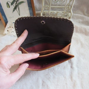 Può includere: Portafoglio in pelle aperto con esterno marrone e interno bordeaux scuro. Il portafoglio ha più scomparti e una chiusura a scatto. I bordi sono cuciti con un motivo decorativo. Il portafoglio è aperto, rivelando l'interno.