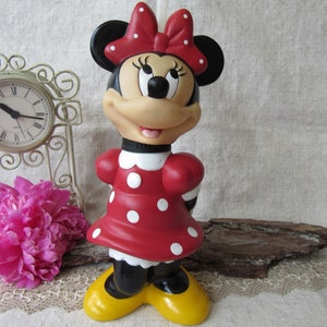 Puede incluir: Una figura de Minnie Mouse, con un vestido rojo con lunares blancos, un lazo a juego y zapatos amarillos. La figura tiene orejas negras y una cara sonriente. Un reloj vintage y flores rosas est&aacute;n en el fondo.