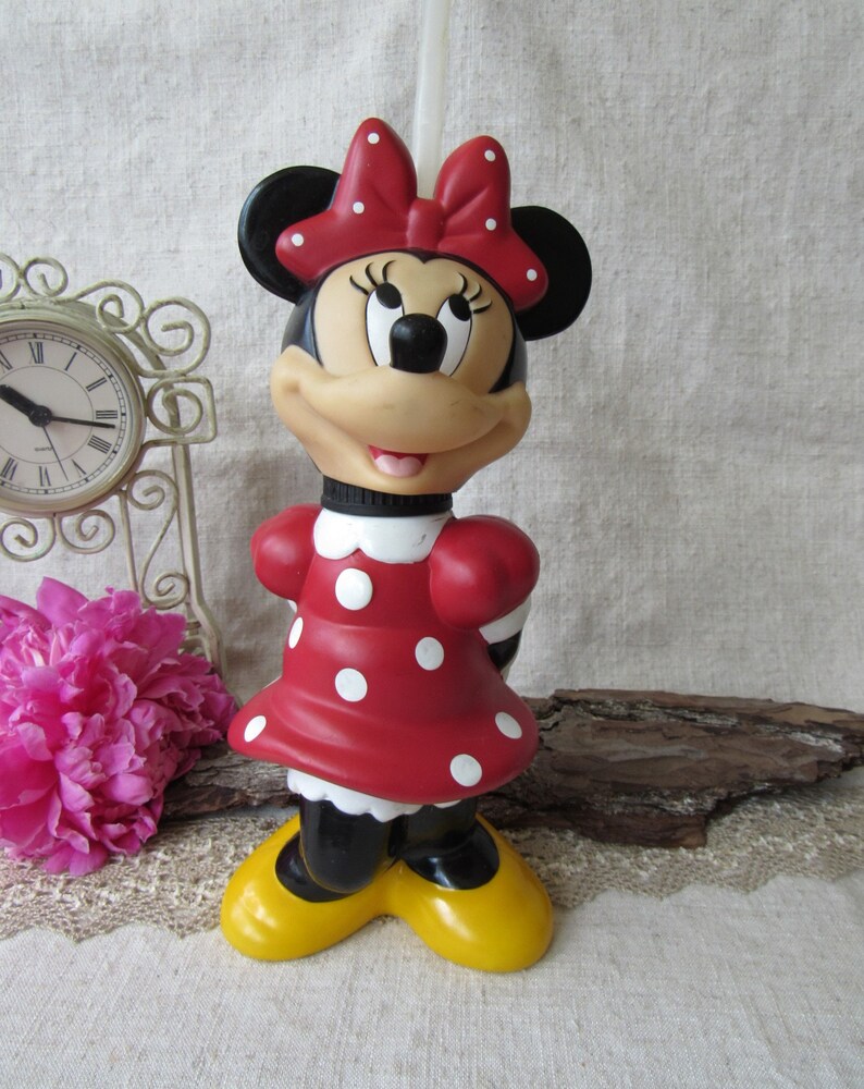 Puede incluir: Una figura de Minnie Mouse, con un vestido rojo con lunares blancos, un lazo a juego y zapatos amarillos. La figura tiene orejas negras y una cara sonriente. Un reloj vintage y flores rosas est&aacute;n en el fondo.