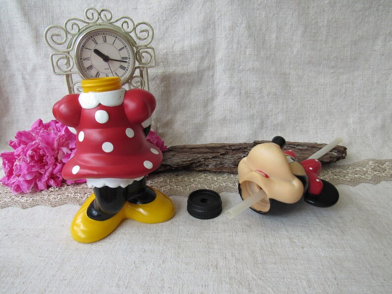 Puede incluir: Una botella de agua con tem&aacute;tica de Minnie Mouse con un vestido rojo con lunares blancos, piernas negras y zapatos amarillos. La botella no tiene cabeza y tiene una tapa negra. Tambi&eacute;n hay una cabeza de Mickey Mouse con una pajita.