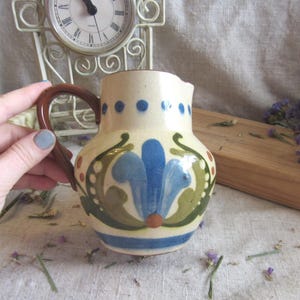 Può includere: Piccolo brocca in ceramica color crema con manico marrone e design floreale blu, verde e arancione. La brocca ha un corpo arrotondato e un bordo leggermente svasato. Punti blu decorano il bordo.