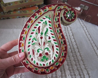 May include: A paisley-shaped decorative box featuring a white center with intricate green and red floral patterns. The border is a deep red with gold-toned accents and small yellow and green teardrop shapes. A circular green floral motif sits at the top.