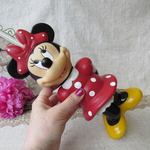 Puede incluir: Una figura de Minnie Mouse con un vestido rojo con lunares blancos y un lazo a juego. La figura tiene zapatos amarillos y orejas negras. La figura est&aacute; sujeta por una mano. Un reloj y flores rosas est&aacute;n en el fondo.