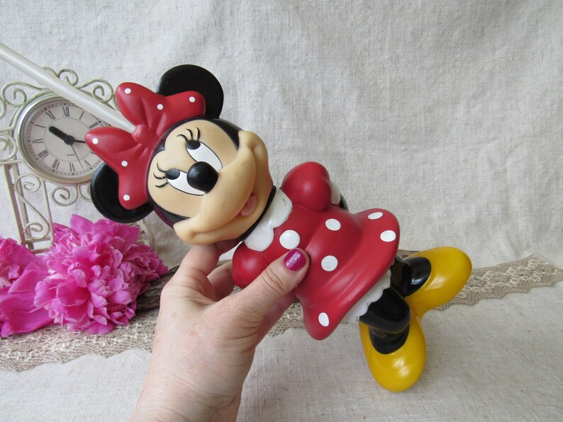 Puede incluir: Una figura de Minnie Mouse con un vestido rojo con lunares blancos y un lazo a juego. La figura tiene zapatos amarillos y orejas negras. La figura est&aacute; sujeta por una mano. Un reloj y flores rosas est&aacute;n en el fondo.