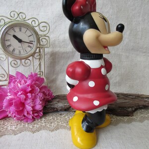 Puede incluir: Una botella de agua con forma de Minnie Mouse. La botella presenta un vestido rojo con lunares blancos, zapatos amarillos y un lazo rojo. La cabeza es negra con una cara bronceada y orejas negras. Una pajita blanca sobresale.