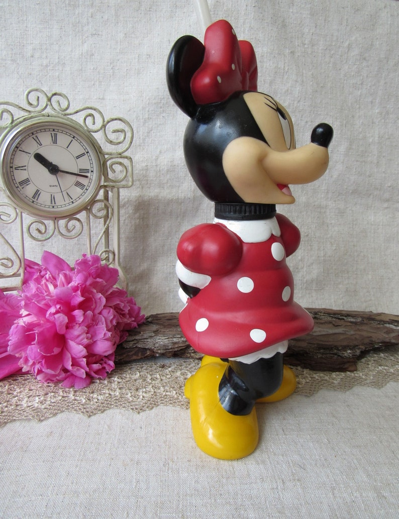 Puede incluir: Una botella de agua con forma de Minnie Mouse. La botella presenta un vestido rojo con lunares blancos, zapatos amarillos y un lazo rojo. La cabeza es negra con una cara bronceada y orejas negras. Una pajita blanca sobresale.