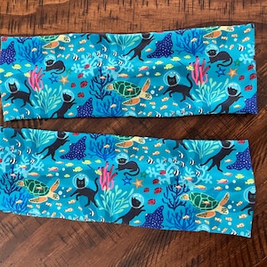 Puede incluir: Dos diademas de tela azul con un patrón repetido de gatos negros con equipo de buceo, tortugas marinas, corales y peces. Las diademas están sobre una superficie de madera.