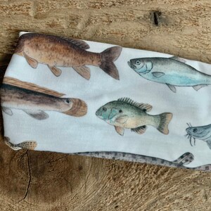 May include: White headband with a colorful fish print. The fish are brown, blue, green, and gray. The headband is made of a soft, stretchy fabric.