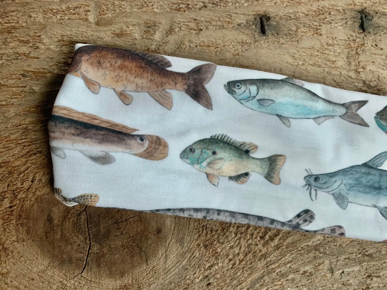 May include: White headband with a colorful fish print. The fish are brown, blue, green, and gray. The headband is made of a soft, stretchy fabric.
