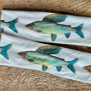 Puede incluir: Dos diademas blancas con un patrón repetido de ilustraciones detalladas de peces. Los peces tienen colores azul, verde y naranja, con aletas y cola grandes. Las diademas están sobre una superficie de madera.