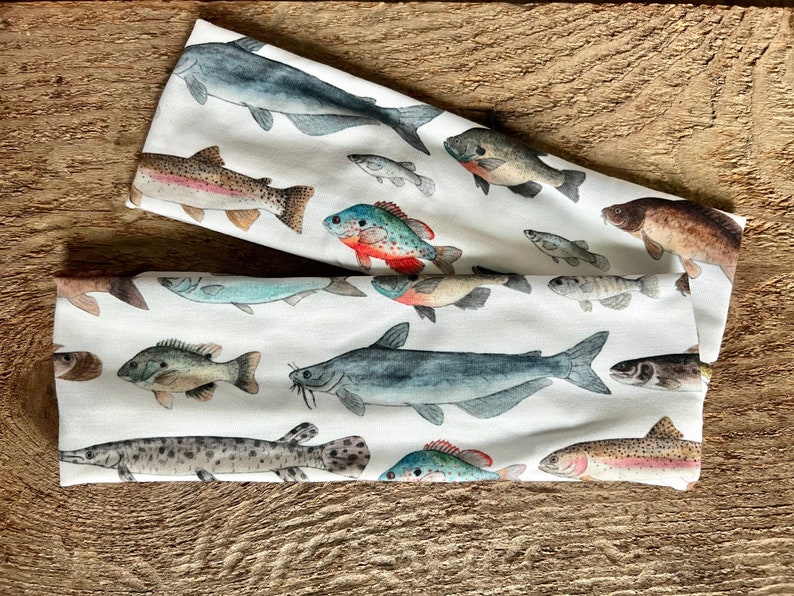 May include: A white fabric headband with a repeating pattern of colorful fish. The fish are illustrated in a watercolor style and include catfish, trout, and bass.