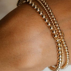 May include: Close-up of three gold beaded bracelets. The bracelets are made of small, round, shiny gold beads. One bracelet has a larger gold ring connecting the strands. The bracelets are worn on a wrist.