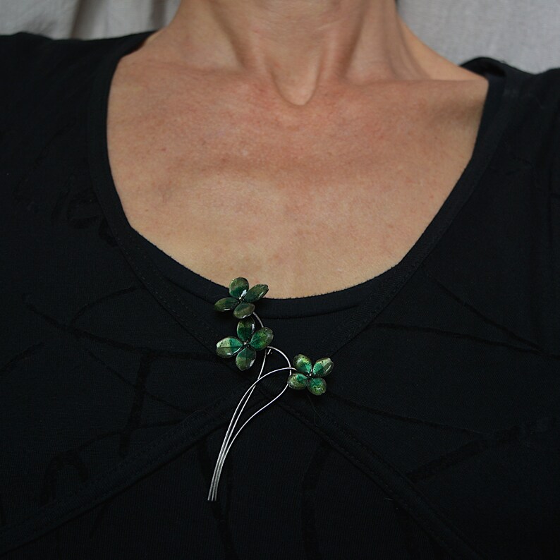 May include: A floral brooch with three clusters of green, gem-like flowers. The brooch has a silver-colored stem and is pinned to a black top. The flowers have a gradient of green hues, creating a shimmering effect.