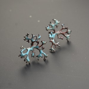 Op de afbeelding: Een paar handgemaakte oorstekers met een uniek, organisch ontwerp. Elke oorbel heeft een vertakte, koraalachtige structuur met een combinatie van turquoise en bruine tinten. De oorbellen zijn geplaatst tegen een donkere achtergrond.
