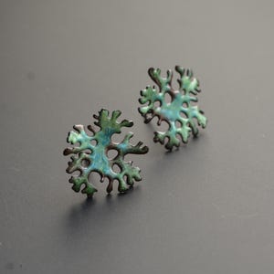 Peut inclure: Une paire de boucles d'oreilles à clous uniques avec un motif de flocon de neige. Les boucles d'oreilles présentent un émail vert et bleu, avec une bordure foncée. Les boucles d'oreilles sont posées sur un fond gris foncé.