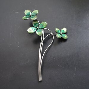 May include: A silver-toned metal brooch featuring a floral design with three clusters of four-leaf clovers. The clover leaves are painted with shades of green and have a textured appearance. The brooch is set against a dark background.