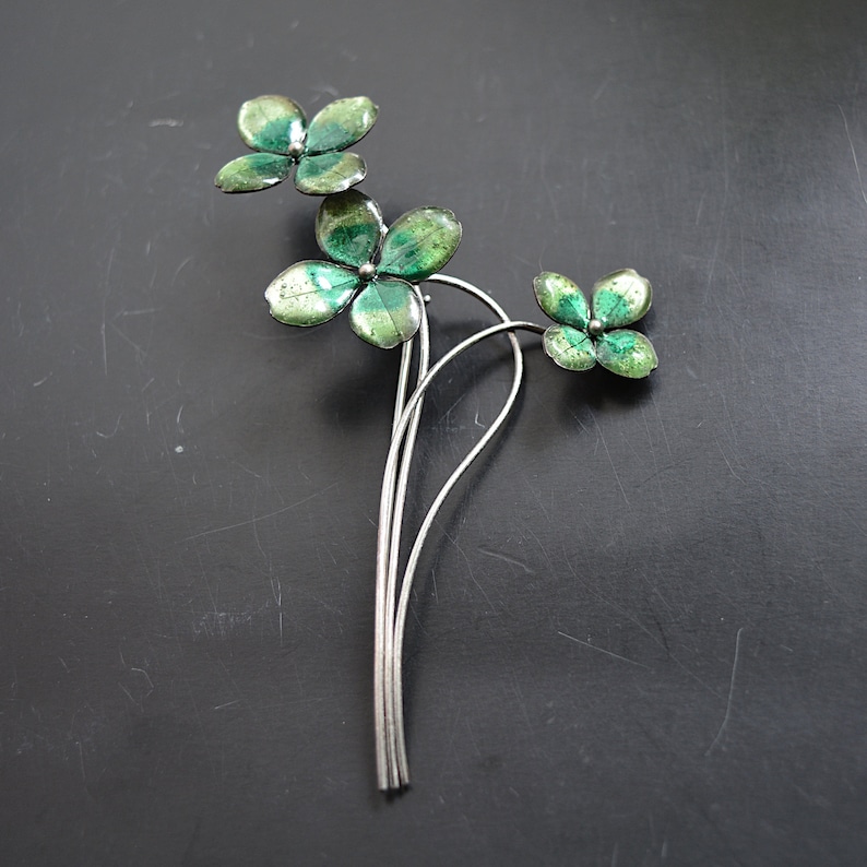 May include: A silver-toned metal brooch featuring a floral design with three clusters of four-leaf clovers. The clover leaves are painted with shades of green and have a textured appearance. The brooch is set against a dark background.
