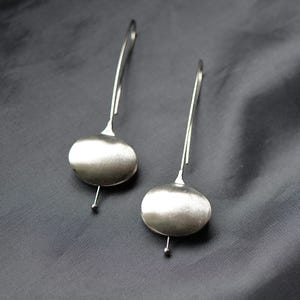 Op de afbeelding: Een paar zilveren oorhangers met een gladde, afgeronde ovale vorm aan de onderkant. De oorhangers hangen aan een dunne zilveren draad.
