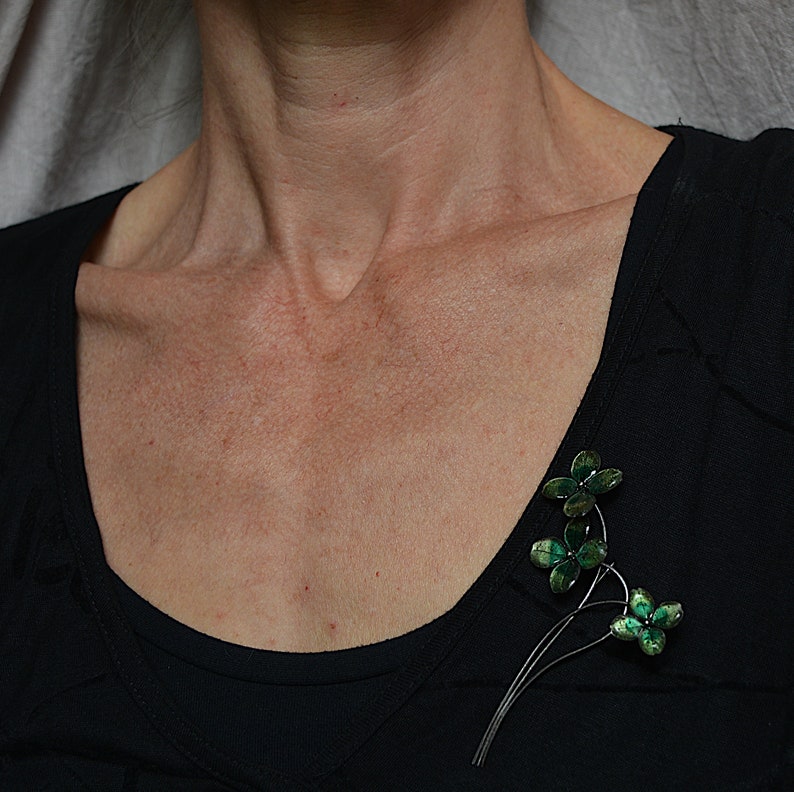 May include: A silver brooch featuring three green clover leaves. The leaves are crafted from metal and have a textured finish. The brooch is pinned to a black shirt.