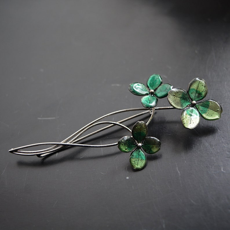 May include: A silver brooch featuring three green enamel flowers. The flowers are clustered together and have a delicate, whimsical design.